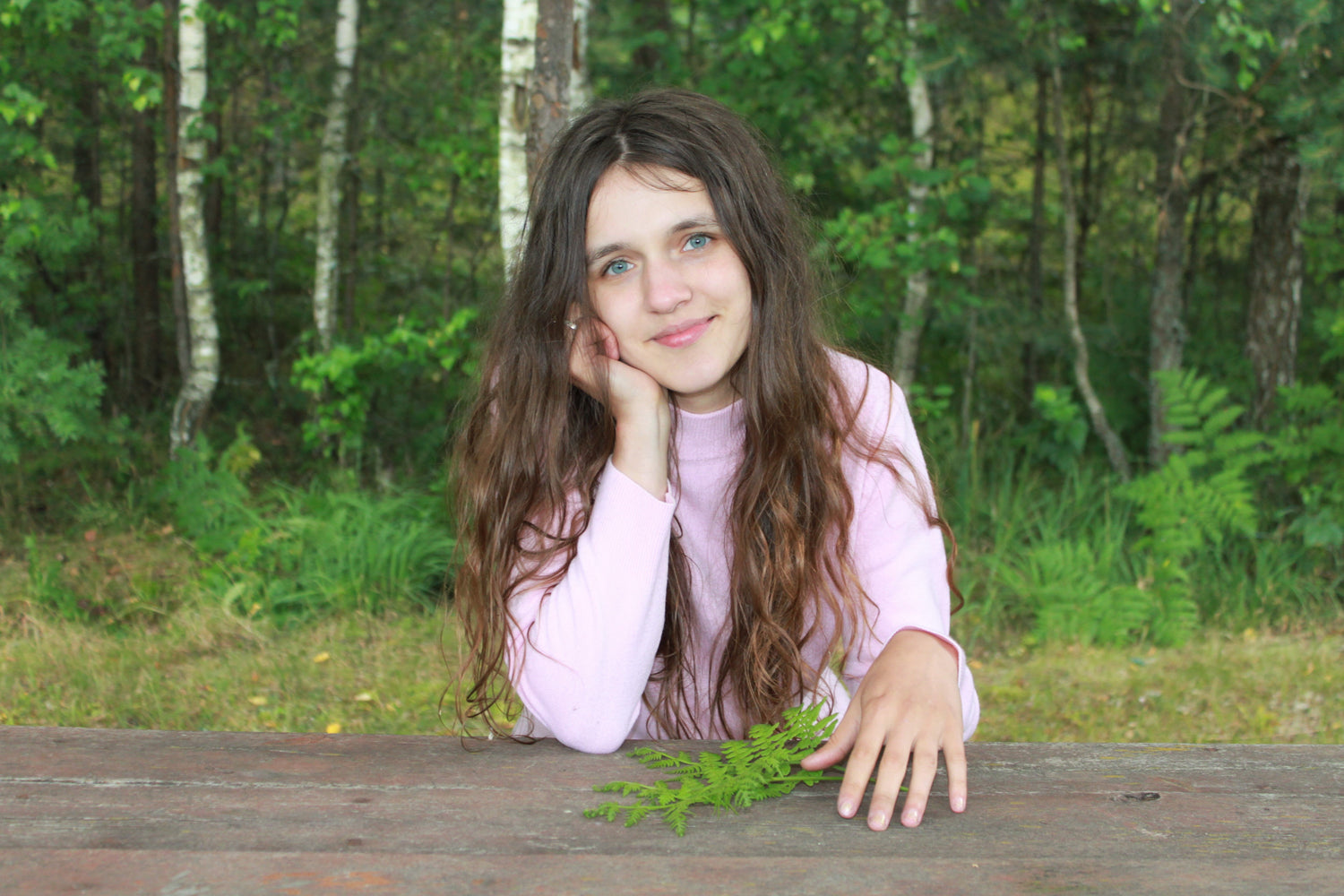 In a pink sweater, Mia is sitting in the forest near the pines, holding a fern, smiling at you! Invites you to enjoy mushroom art!